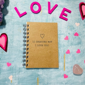 May include: A brown notebook with a spiral binding and the text "10 reasons why I love you" on the cover. The notebook is surrounded by pink felt hearts and the word "LOVE" spelled out in pink felt letters.