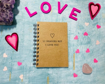 May include: A brown notebook with a spiral binding and the text "10 reasons why I love you" on the cover. The notebook is surrounded by pink felt hearts and the word "LOVE" spelled out in pink felt letters.