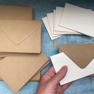 以下が含まれることがあります： クラフト紙の封筒と白いカード。茶色の封筒がいくつか青い面に配置されています。手は白いカードを持っており、茶色の封筒の中に収まる様子を示しています。封筒は正方形です。