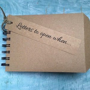 May include: A brown notebook with a spiral binding and a brown paper tag attached. The tag has the text "Letters to open when..." written on it.