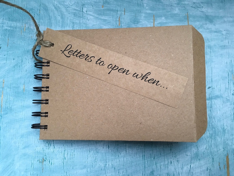May include: A brown notebook with a spiral binding and a brown paper tag attached. The tag has the text "Letters to open when..." written on it.