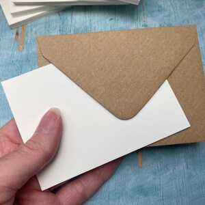 以下が含まれることがあります： 白いカードが茶色のクラフト紙の封筒に部分的に挿入されています。カードは手で持たれています。同様のカードの束が背景に見えます。