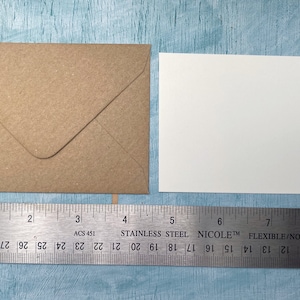 以下が含まれることがあります： クラフト紙の封筒と白いカードが、ステンレス製の定規の隣に表示されています。定規はセンチメートル単位の測定値を表示しており、封筒の幅は約10cmです。カードは正方形です。