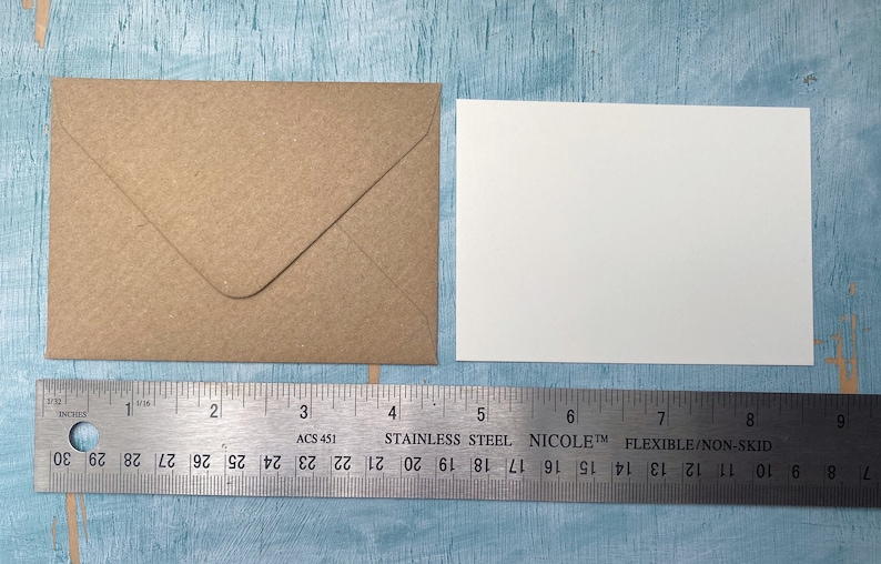 以下が含まれることがあります： クラフト紙の封筒と白いカードが、ステンレス製の定規の隣に表示されています。定規はセンチメートル単位の測定値を表示しており、封筒の幅は約10cmです。カードは正方形です。