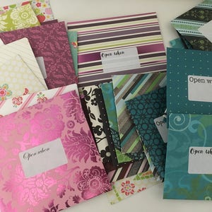May include: A collection of decorative envelopes with various patterns and colors, including floral, striped, and geometric designs. Each envelope has a label that reads "Open when". The envelopes are in shades of pink, green, purple, and teal.