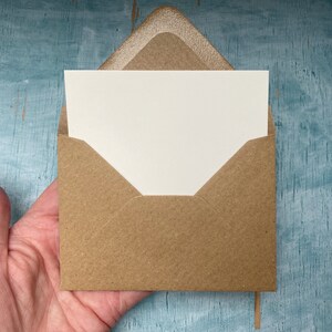 以下が含まれることがあります： クリーム色のカードが入った、小さな正方形のクラフト紙の封筒。封筒は開いており、カードが見えています。背景は、質感のあるターコイズブルーの表面です。