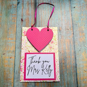 May include: A pink heart-shaped tag with a textured surface and a pink ribbon. The tag is attached to a card with a floral pattern and the words "Thank you Mrs Kelly" in black script.