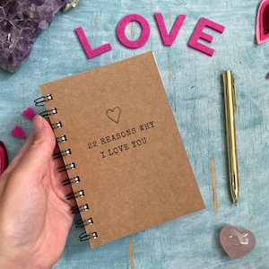May include: A brown notebook with a spiral binding. The notebook has the text "22 Reasons Why I Love You" printed on the front cover. A hand is holding the notebook. There are pink felt hearts and a pink crystal on a blue background.
