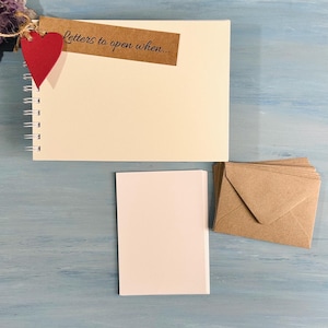 May include: A blank notebook with a brown tag that says "letters to open when..." and a red heart attached. There are also blank white cards and brown envelopes on a blue wooden surface.