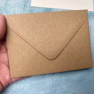 以下が含まれることがあります： 手で持たれた、尖ったフラップが付いた小さな茶色のクラフト紙封筒。封筒は、明るい青色のテクスチャ加工された表面にあります。背景には白いカードが見えます。