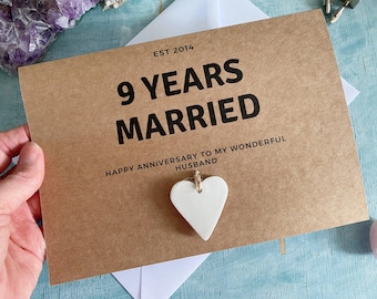 Könnte beinhalten: Eine braune Karte mit dem Text "9 YEARS MARRIED" in Schwarz. Die Karte trägt auch den Text "EST 2014" und "HAPPY ANNIVERSARY TO MY WONDERFUL HUSBAND". Ein weißes herzförmiges Ornament hängt an der Karte.