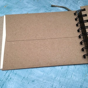May include: A brown kraft paper notebook with a white spine and a black wire-o binding. The notebook is closed and lying on a blue surface.