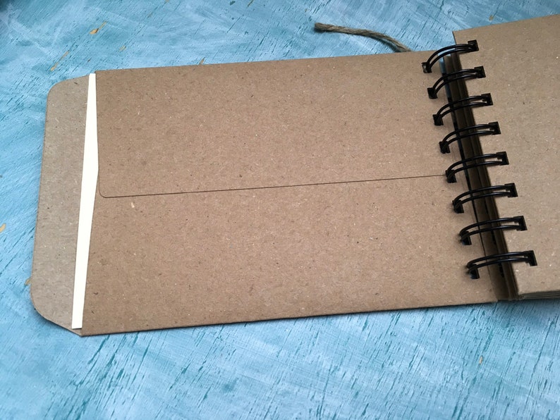 May include: A brown kraft paper notebook with a white spine and a black wire-o binding. The notebook is closed and lying on a blue surface.
