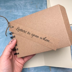 May include: A brown kraft paper journal with a spiral binding and a brown paper tag that says "Letters to open when..." The journal is tied with twine.