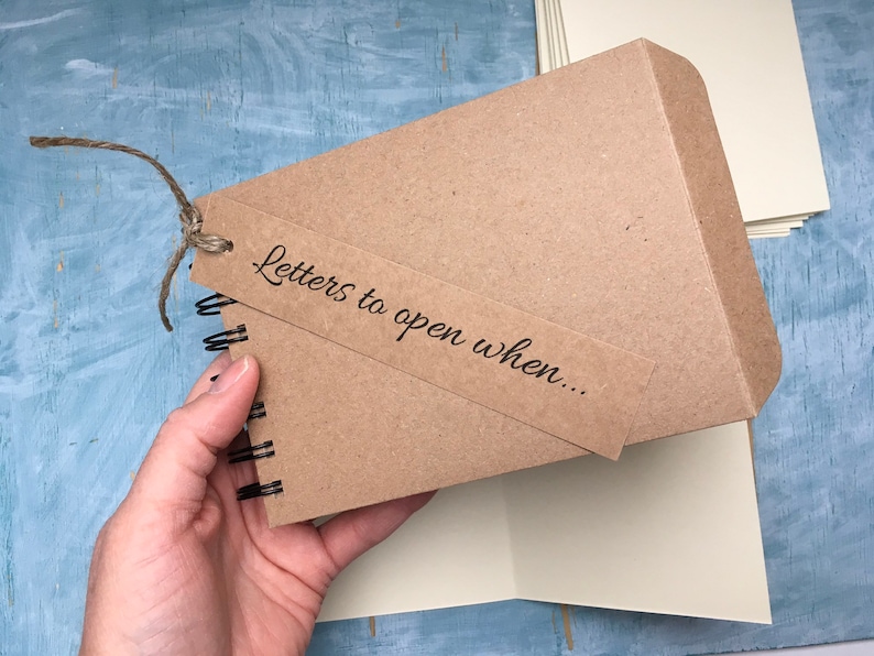 May include: A brown kraft paper journal with a spiral binding and a brown paper tag that says "Letters to open when..." The journal is tied with twine.