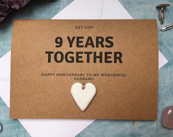 May include: A brown card with the text "9 YEARS TOGETHER" and "EST 2011". Below the text is "HAPPY ANNIVERSARY TO MY WONDERFUL HUSBAND". A white heart-shaped ornament hangs from the card. The card is on a white envelope.