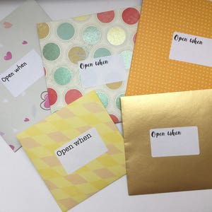 May include: A collection of colorful envelopes with the text "Open when" on white labels. The envelopes feature various patterns, including hearts, polka dots, and geometric designs. The envelopes are in shades of pink, yellow, gold, and green.