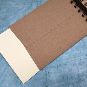 May include: A brown notebook with a spiral binding and a blank white page sticking out. The notebook is on a blue wooden surface.