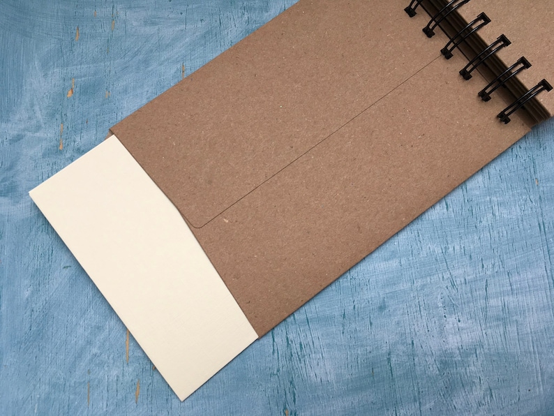 May include: A brown notebook with a spiral binding and a blank white page sticking out. The notebook is on a blue wooden surface.