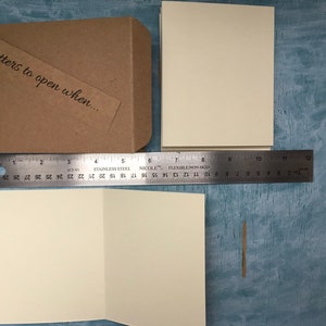 May include: A brown kraft paper journal with a tag that says "Letters to open when..." and a spiral binding. The journal is next to a blank white card and a ruler.