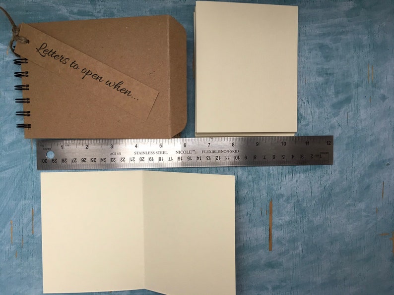 May include: A brown kraft paper journal with a tag that says "Letters to open when..." and a spiral binding. The journal is next to a blank white card and a ruler.