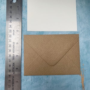 以下が含まれることがあります： 白い長方形のカードと茶色の封筒が定規の隣に表示されています。定規はインチ単位の測定値を表示します。カードの幅は約10cmです。封筒は折りたたまれ、封印されています。