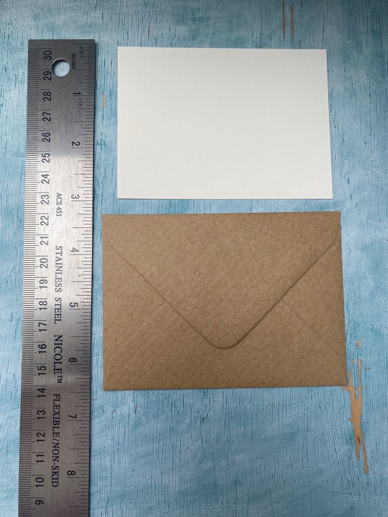 以下が含まれることがあります： 白い長方形のカードと茶色の封筒が定規の隣に表示されています。定規はインチ単位の測定値を表示します。カードの幅は約10cmです。封筒は折りたたまれ、封印されています。