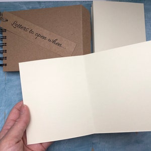 May include: A brown spiral bound notebook with a tag that says "Letters to open when..." The notebook is open to reveal blank white pages.