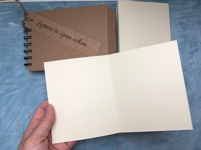 May include: A brown spiral bound notebook with a tag that says "Letters to open when..." The notebook is open to reveal blank white pages.