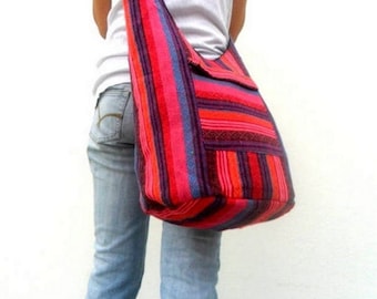 Può includere: Una grande borsa a tracolla a righe colorate con una lunga tracolla indossata sulla spalla. La borsa è realizzata in tessuto intrecciato e presenta una varietà di colori, tra cui rosso, rosa, viola e blu.