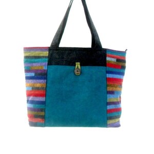 May include: A teal blue tote bag with black straps and colourful striped fabric on the sides. The bag has a flap with a wooden button closure.