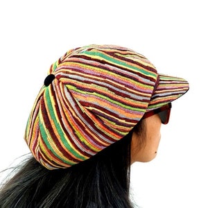 Pode incluir: Um boné de estilo newsboy com listras coloridas e um botão preto no topo. O boné tem uma aba que se curva para baixo nas laterais e uma parte superior plana.