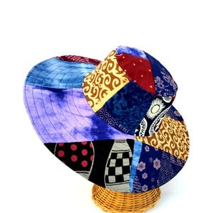 Puede incluir: Un sombrero de sol de ala ancha hecho de tela de patchwork en tonos de azul, morado, rojo, amarillo y marrón. El sombrero tiene un estilo único y bohemio.