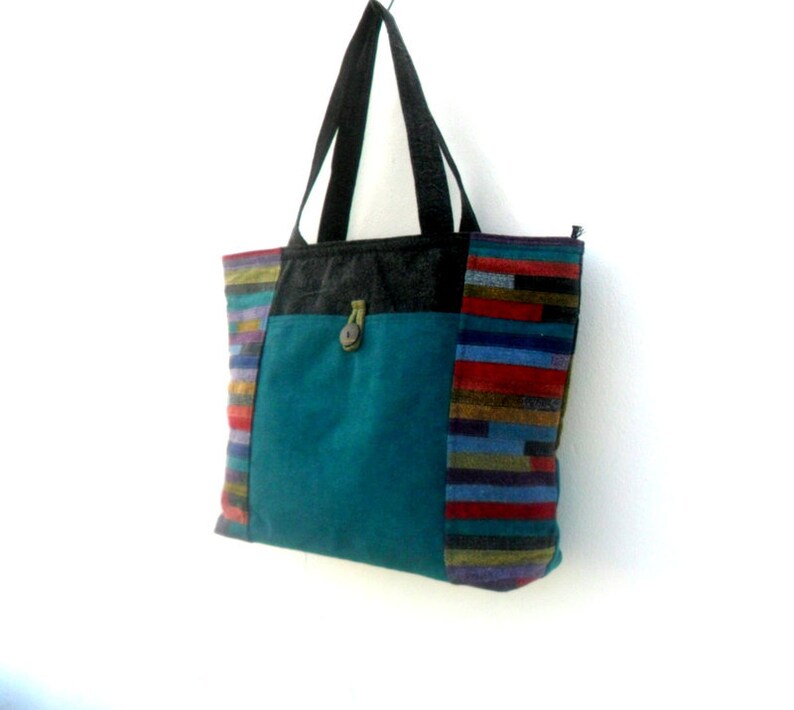 May include: A teal blue tote bag with a black handle and a colourful striped panel on the side. The bag has a flap with a button closure.