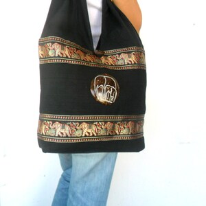 May include: Black cotton tote bag with a decorative elephant design. The bag has a brown and gold trim with elephants and floral patterns. The bag has a round, brown, and white elephant button.