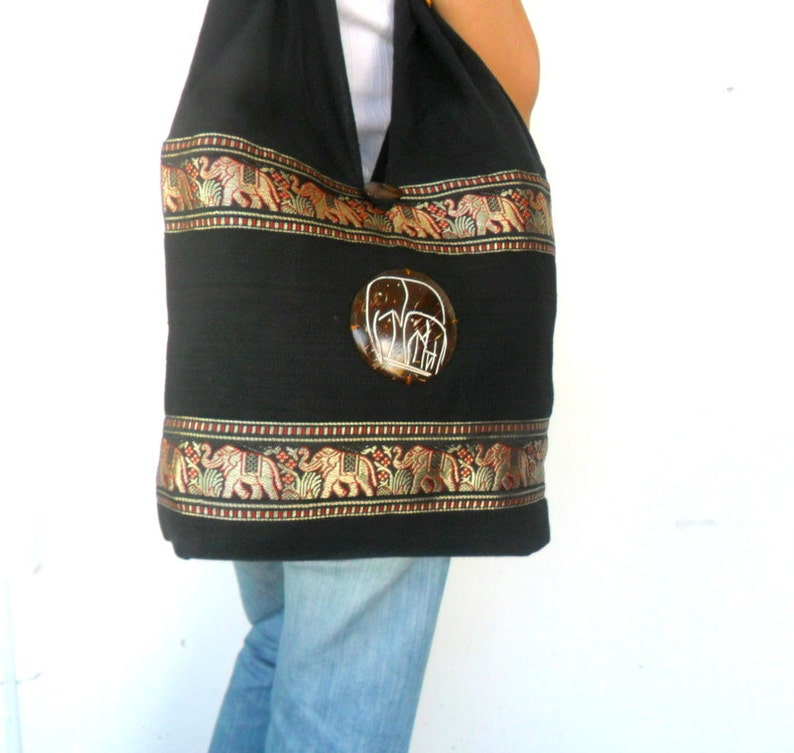 May include: Black cotton tote bag with a decorative elephant design. The bag has a brown and gold trim with elephants and floral patterns. The bag has a round, brown, and white elephant button.