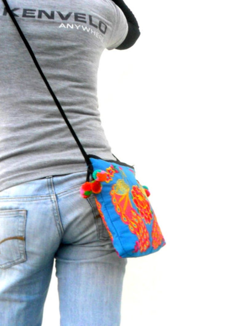 May include: A person wearing blue jeans and a grey t-shirt with the text "Kenvelo Anywhere" on the front. The person is wearing a blue and pink floral crossbody bag with a black strap and colorful tassels.