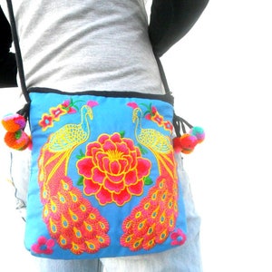 May include: A blue fabric crossbody bag with embroidered peacocks and a pink flower. The bag has a black strap and colorful pom-poms on the ends.