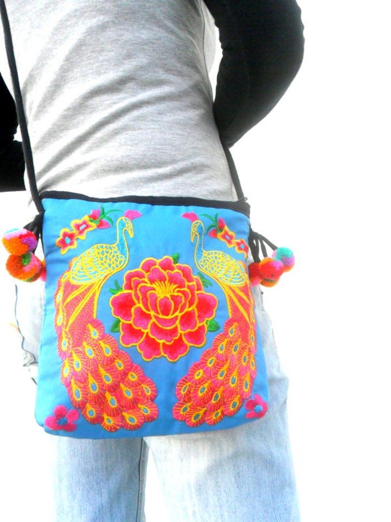 May include: A blue fabric crossbody bag with embroidered peacocks and a pink flower. The bag has a black strap and colorful pom-poms on the ends.