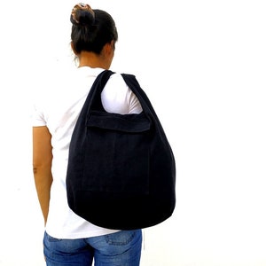 Pode incluir: Uma bolsa tote de lona preta com um bolso grande na frente. A bolsa &eacute; usada no ombro de uma pessoa que usa uma camiseta branca e jeans azul.