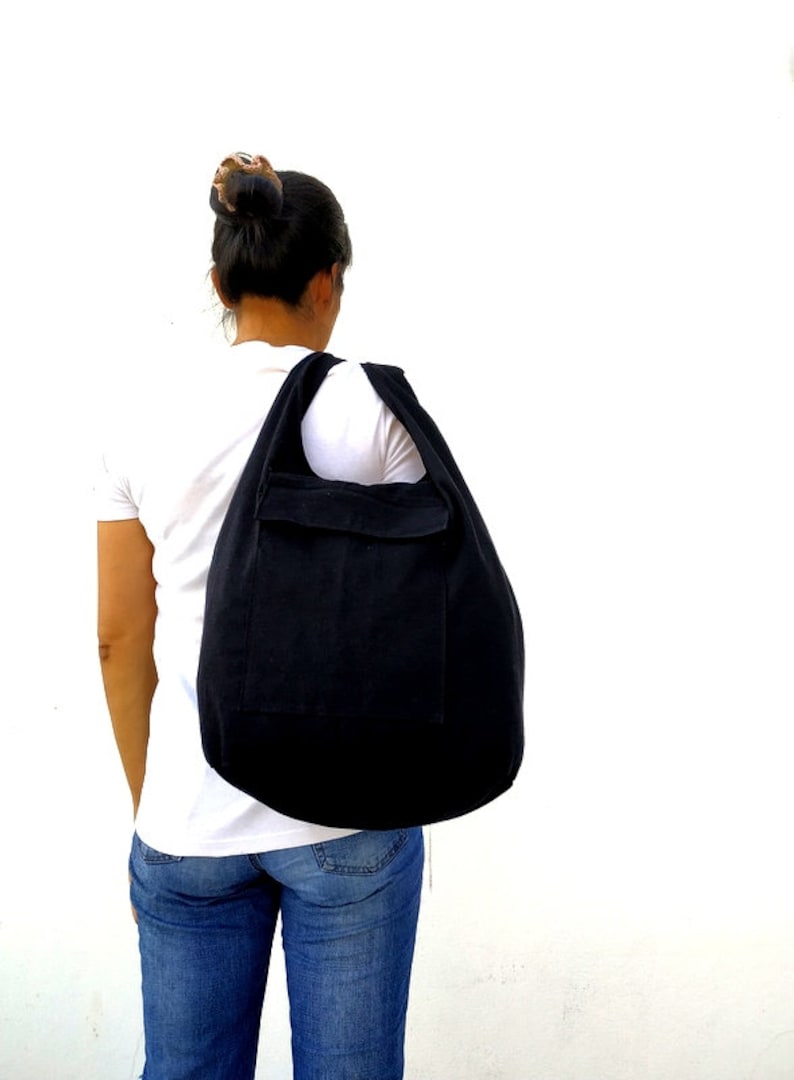 Pode incluir: Uma bolsa tote de lona preta com um bolso grande na frente. A bolsa &eacute; usada no ombro de uma pessoa que usa uma camiseta branca e jeans azul.