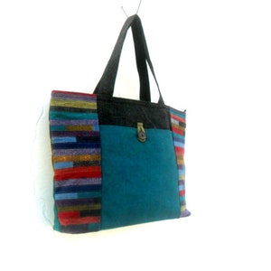 May include: A teal blue tote bag with a black and grey fabric pocket on the front. The bag has a colourful striped fabric trim on the sides.