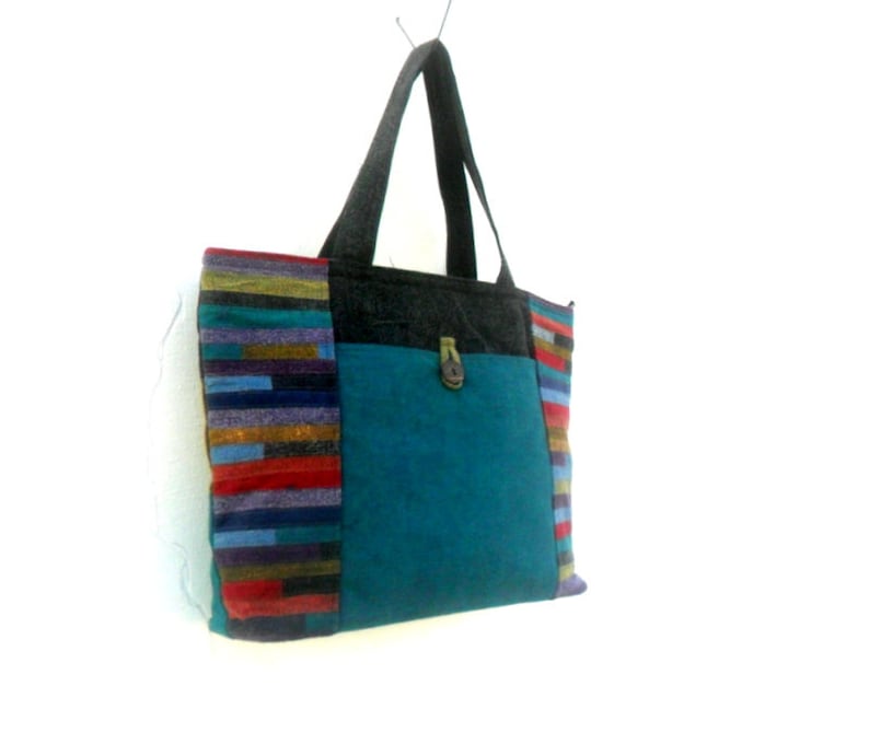 May include: A teal blue tote bag with a black and grey fabric pocket on the front. The bag has a colourful striped fabric trim on the sides.