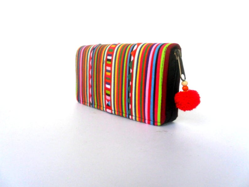 Puede incluir: Una cartera con cremallera de rayas coloridas con un abalorio de pomp&oacute;n rojo. La cartera est&aacute; hecha de tela y tiene una cremallera negra.