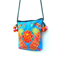 May include: A small blue crossbody bag with a floral and peacock design. The bag has a black strap and two fluffy orange and black pom-poms.