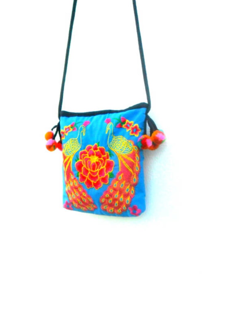 May include: A small blue crossbody bag with a floral and peacock design. The bag has a black strap and two fluffy orange and black pom-poms.