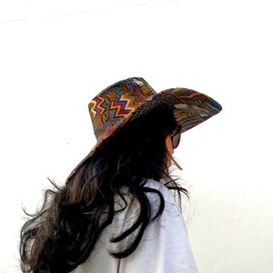 Puede incluir: Una mujer con el pelo largo y oscuro lleva un sombrero de ala ancha con un estampado geom&eacute;trico colorido. El sombrero est&aacute; hecho de tela y tiene un ala ligeramente curvada. La mujer est&aacute; de espaldas a la c&aacute;mara y mira hacia la derecha.