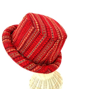 以下が含まれることがあります： 幅広のつばが付いた赤と金の模様のバケットハット。帽子は織り生地で作られており、わずかに丸みを帯びたクラウンがあります。