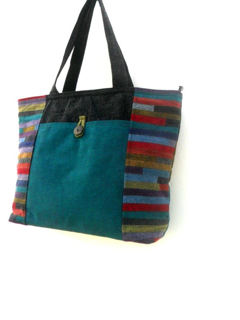 May include: A teal blue tote bag with a black zipper and a striped fabric panel on the side. The bag has a black handle and a small pocket on the front with a wooden button.