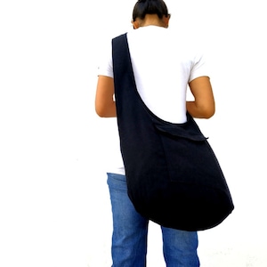 Pode incluir: Uma pessoa vestindo jeans azul e uma camiseta branca est&aacute; carregando uma grande bolsa tote de lona preta no ombro.